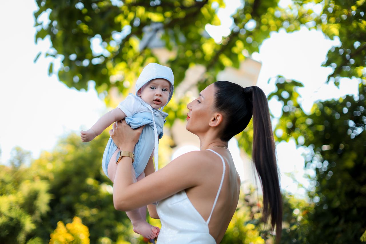 why-choose-us A joyful mother holding her baby under a green leafy tree on a sunny day.