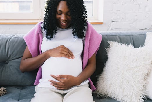 pexels photo 6991886 6991886 Cheerful pregnant African American female touching tummy gently while sitting on comfortable sofa with pillows in light room at home